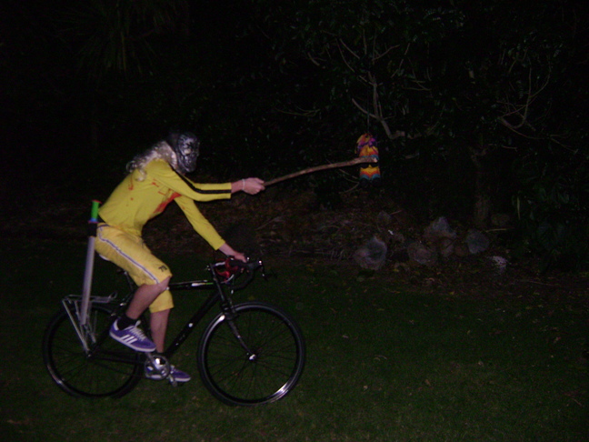 Right side view of a person riding a bike, wearing a yellow costume, striking a piñata, on grass at night 
